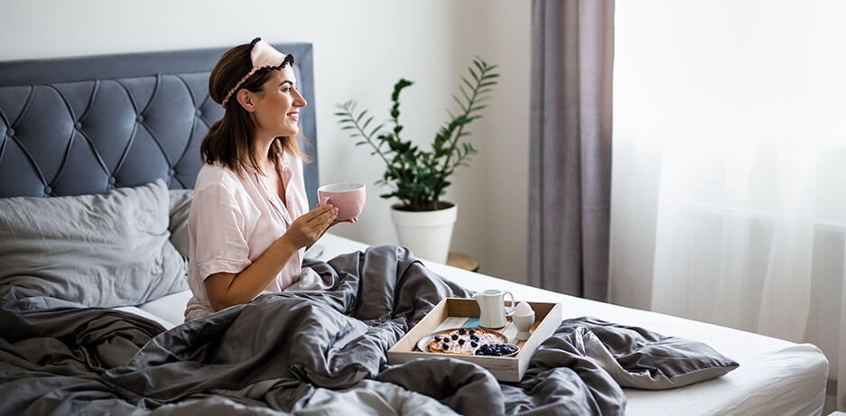 Brunette lady having breakfast in a bed with a grey headboard and grey and white bedding